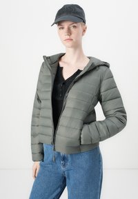 Grønn pufferjakke med glidelås, hette og horisontal quiltedesign. Kombinert med en svart henley-genser og blå jeans. Svart caps på hodet.