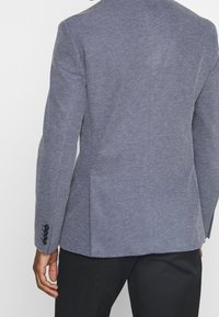 Man i en strukturert blå kavaj med svarta knappar på ärmarna och svarta byxor, visad bakifrån och från midjan ner.