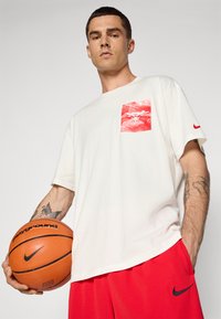 Hvid bomuld t-shirt med en rød trykt lomme, der har et bælg-logo. Korte ærmer, båret med røde shorts. Hånd der holder en basketball.