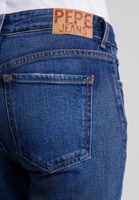 Bakvy av blå denimjeans med en brun läderlapp med bleknad röd "Pepe Jeans" text ovanför den högra bakfickan.