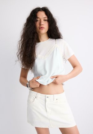 Ung kvinde med langt krøllet hår løfter en hvid T-shirt og viser maven, iført en ærmeløs top med blondekanter ovenpå og et hvidt denimskørt.