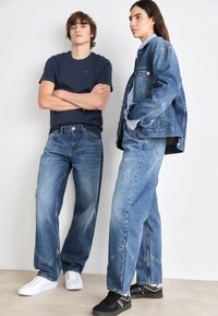 To modeller står side om side, iført blå denimjeans og casual skjorter. Den mannlige modellen har på seg en marineblå T-skjorte; den kvinnelige modellen har på seg en denimjakke.