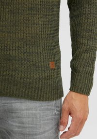 Strukturierter grüner Pullover mit Rippdesign, versehen mit einem braunen Lederlogo-Patch am Saum, kombiniert mit hellgrauen Hosen.