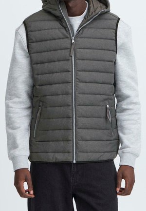 Uomo che indossa un gilet trapuntato grigio con cappuccio sopra una felpa grigio chiaro e pantaloni neri, in piedi con le braccia rilassate lungo i fianchi.