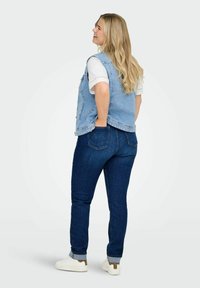 Besidėjusi moteris su ilgu šviesiais plaukais, vilkinti džinsinę liemenę, baltą marškinėlį, tamsiai mėlynus džinsus ir baltus batelius su leopardų raštu.