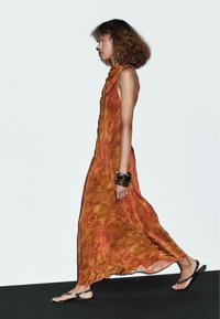 Robe maxi orange avec motif orné, sans manches, en tissu fluide et fente sur le côté, associée à des sandales noires et des bracelets empilés.