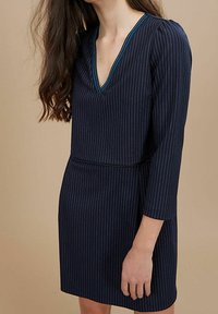 Marineblaues Kleid mit vertikalen weißen Streifen, V-Ausschnitt, Dreiviertelärmeln und taillierter Taille. Akzentuiert mit türkisfarbenen Nähten entlang des Ausschnitts.