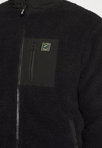 Chaqueta de forro polar negra que presenta un panel de tela negra lisa y un bolsillo en el pecho con cremallera y un parche de logo verde. Superficie texturizada en toda la prenda.