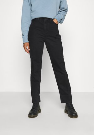 Personne portant un jean noir taille haute à jambes droites, des bottes noires à lacets et un sweat-shirt bleu clair à manches longues. Une main dans la poche.