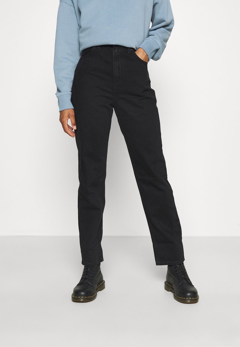 Personne portant un jean noir taille haute à jambes droites, des bottes noires à lacets et un sweat-shirt bleu clair à manches longues. Une main dans la poche.