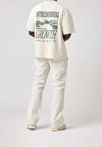 T-shirt crème à manches courtes avec un motif imprimé dans le dos, associé à un pantalon cargo beige clair avec de multiples poches et une coupe décontractée.