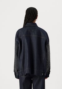Chaqueta de mezclilla oscura con acentos de tachuelas en las mangas, con un corte holgado y detalles de costura visibles. Logotipo en la parte posterior visible en el collar.