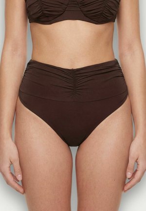 Mujer con braguitas de bikini marrones de tiro alto con fruncido en el centro delantero y las caderas, visibles desde el abdomen hasta la parte superior de los muslos.