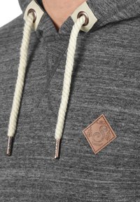 Hoodie gris avec un tissu texturé, doté d'un cordon en rope beige et d'une patch logo en cuir marron sur la poitrine. Accents en métal argenté.