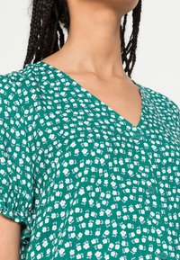 Blusa floreale verde con scollatura a V, maniche corte e piccoli fiori bianchi con accenti verdi. Include un dettaglio con bottoni sul davanti.