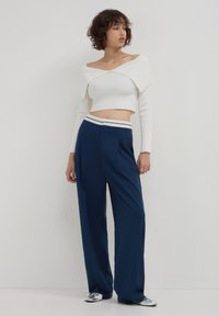 Jeune femme portant un top court côtelé blanc sans manches, un pantalon large marine avec une ceinture blanche, et des chaussures argentées à bout pointu, se tenant devant un mur blanc.