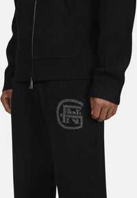 Czarny sweter zapinany na zamek z dzianiny, z prążkowanymi mankietami, w zestawie z pasującymi czarnymi dresami z szarym nadrukiem logo na udzie. Miękka faktura.