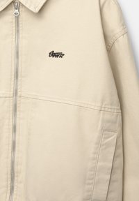 Veste beige avec fermeture éclair sur le devant, col, poche latérale et petit logo noir brodé sur la zone gauche de la poitrine.
