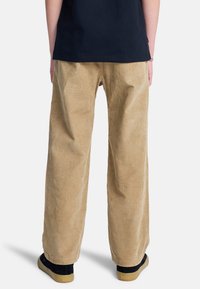 Personne portant un pantalon en velours côtelé beige ample, une chemise noire et des baskets noires à semelles en gomme, debout de dos sur un fond blanc.