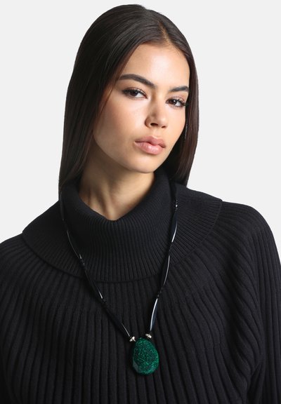 Jeune femme aux cheveux raides et foncés porte un pull à col roulé noir côtelé et un collier noir avec un pendentif en pierre verte de grande taille.