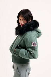 Zelená bomber bunda s čiernou umelou kožušinou na kapucni, rebrovými manžetami a ružovou nášivkou na rukáve, s detailom zipsového vrecka.