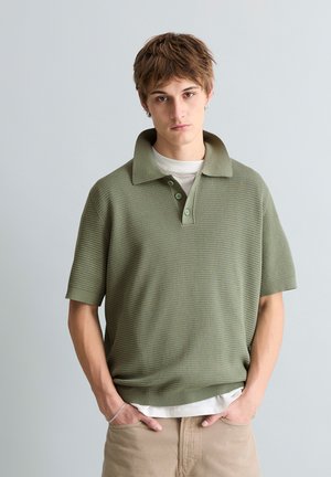 Jeune personne aux cheveux bruns courts portant un polo texturé vert olive clair à manches courtes sur une chemise blanche et un pantalon beige, les mains dans les poches.