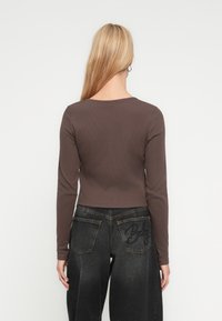 Ξανθιά γυναίκα με ίσια μαλλιά φοράει καφέ εφαρμοστή μπλούζα με μακριά μανίκια και μαύρο τζιν με κεντημένες τσέπες στην πλάτη, στέκεται μπροστά σε λευκό φόντο.