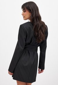 Robe blazer noire à rayures verticales avec des manches longues, une taille ceinturée, une texture lisse et un design ajusté. Motif de rayures verticales distinctif.