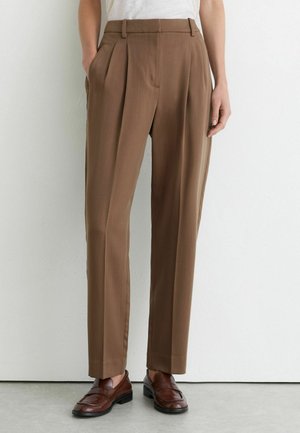 Person trägt hoch taillierte braune maßgeschneiderte Hose mit Falten, Seitentaschen und dunkelbraunen Lederloafers und steht an einer weißen Wand.