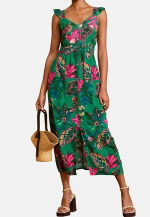 Woman in a green sleeveless floral dress holding a straw handbag, wearing beige high-heeled sandals, standing against a white background.