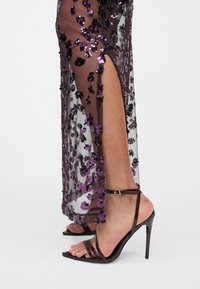 Jambes dans une robe violette transparente avec des sequins et une fente haute, portant des sandales à talons hauts à lanières violettes foncées sur un fond blanc.
