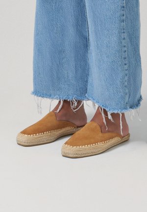 Nohy v hnědých semišových espadrilách na zavlékání s pletenými podrážkami, v kombinaci s světle modrými džíny s roztrhaným lemem na neutrální podlaze.