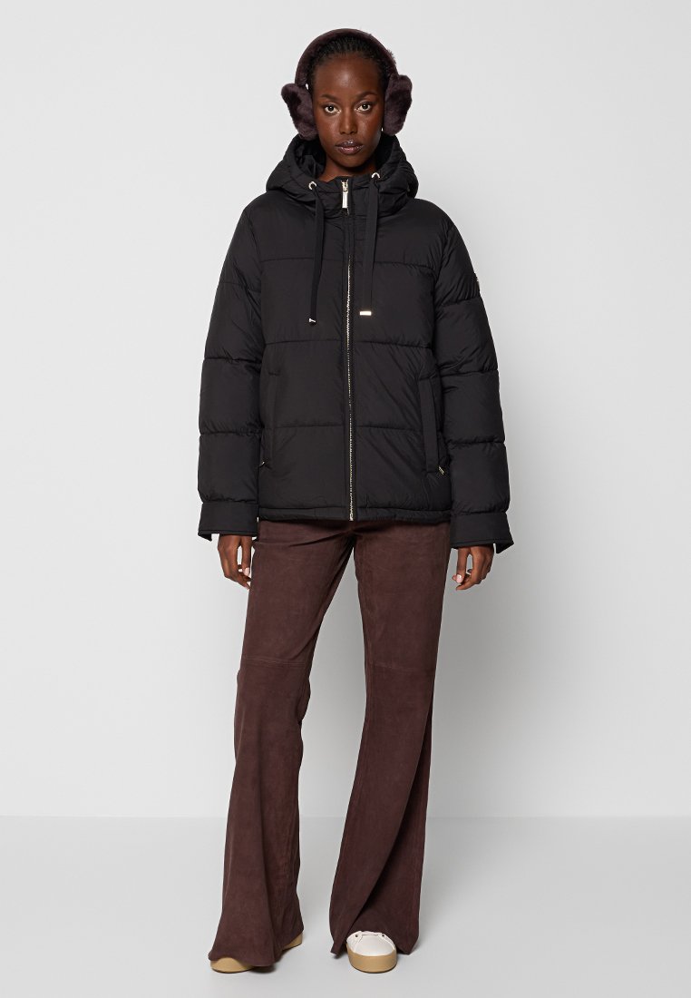 Chaqueta acolchada negra con capucha, cuenta con cierre de cremallera y cordones. El modelo lleva pantalones acampanados marrones y orejeras de piel. Textura de tejido suave.