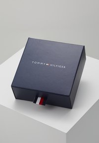 Temno plava kutija s glatkom teksturom i gornjim logotipom koji glasi "TOMMY HILFIGER." Sadrži mali povlačni trak u crvenoj, bijeloj i plavoj boji.