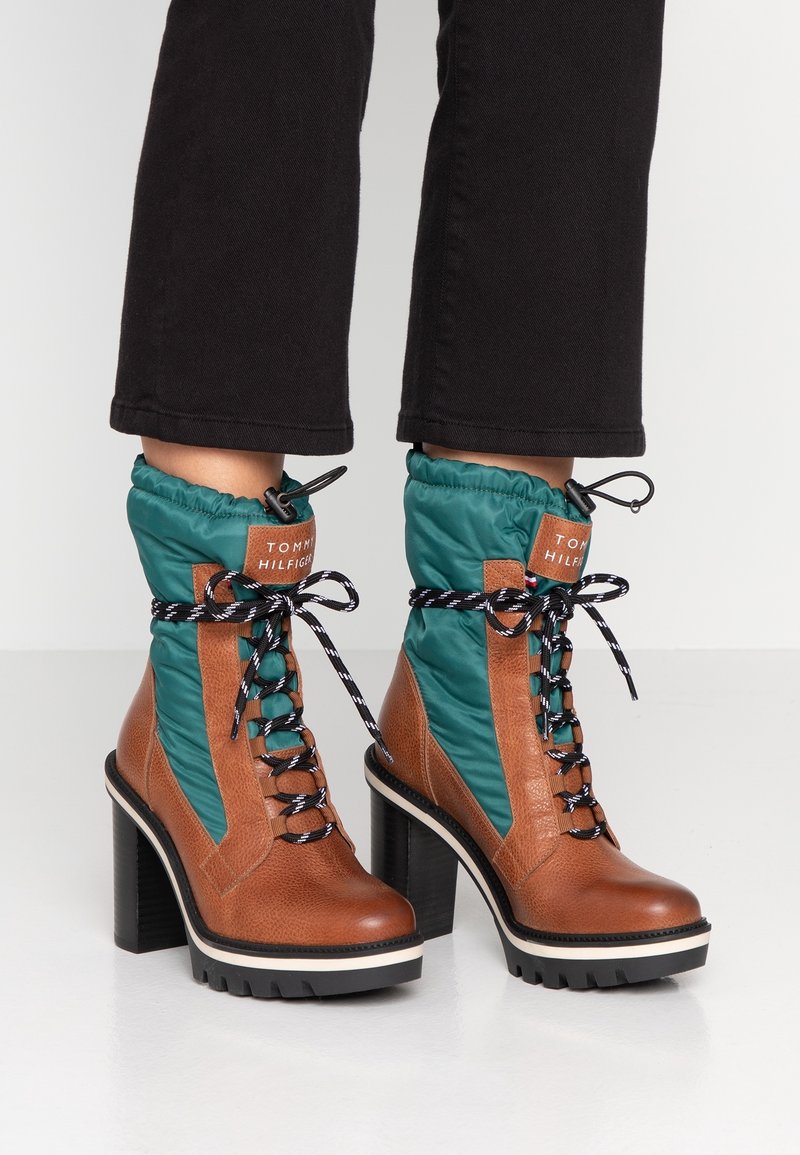 Bottes chevilles en cuir marron et tissu vert avec lacets noirs et blancs, talons hauts épais, portées avec un pantalon noir court.