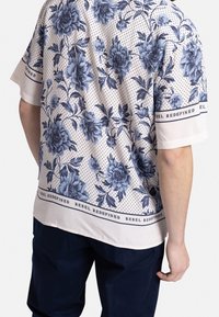 Man in een kortemouwen shirt met een blauw bloemenpatroon en kleine zwarte stippen op een beige achtergrond, gecombineerd met donkere broek.