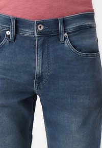 Temno modri kavbojke zobe dvema tradicionalnima žepoma, kontrastnim šiviljernim šivom ter gumbom za zapiranje s sistemom zadrge.