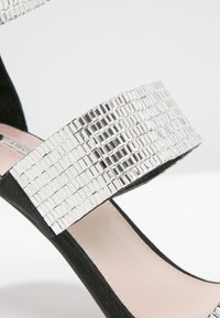 Sandale en daim noir avec une large bride argentée miroir présentant un motif rectangulaire, un design à bout ouvert et un intérieur rose pâle.