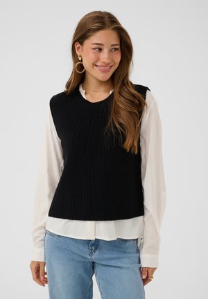Joven mujer con cabello largo y castaño sonriendo, lleva grandes pendientes de aro, un suéter negro sin mangas sobre una blusa blanca y vaqueros azul claro.