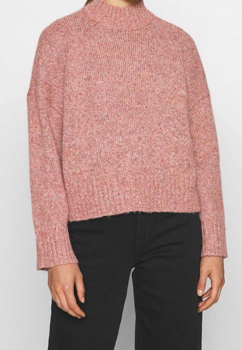 Pull tricoté de couleur rose avec un col montant, une texture côtelée, des épaules tombantes et une coupe courte. Associé à un pantalon foncé.
