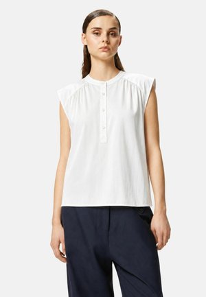 Donna che indossa una camicetta bianca senza maniche con bottoni frontali e spalle strutturate, abbinata a pantaloni larghi blu navy scuro, in piedi dritta.