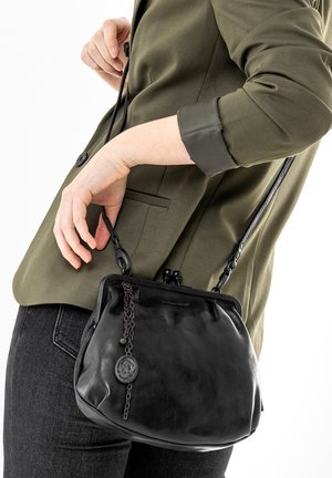 Sac à main en cuir noir de forme arrondie, avec des détails de chaîne et une fermeture à clip. Porté sur une veste olive, avec de longues anses.