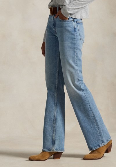 Személy világoskék, egyenes szárú farmernadrágot, barna bőr övet, szürke hosszú ujjú felsőt és világosbarna, velúr magas sarkú bokacsizmát visel, semleges padlón áll.