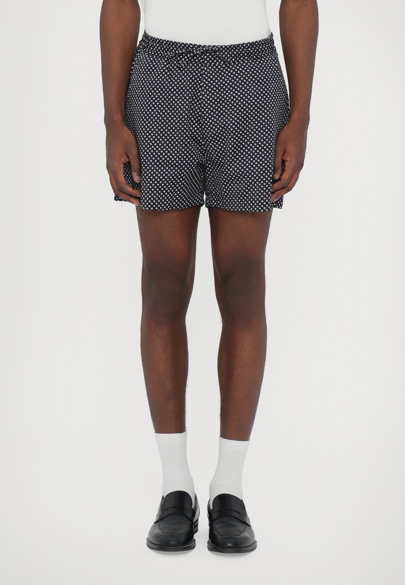 Shorts noirs avec un motif à pois blancs, taille élastique et fentes sur les côtés, associés à des chaussettes blanches et des mocassins noirs.