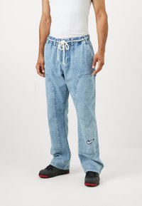 Világoskék farmernadrág laza fazonnal, rugalmas derékkal és húzózsinórral, oldalsó zsebekkel és egy kis fekete Nike logóval a bal lábon.