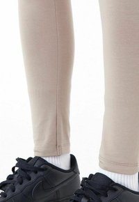 Leggings ajustés beiges en tissu lisse, associés à des baskets noires Nike et à des chaussettes blanches mi-mollet. Coutures et textures minimales visibles.