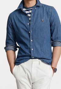 Jeansskjorta med strukturerad krage, kontrasterande blå och vit randig undertröja, ihop med vita byxor och en brun klocka.