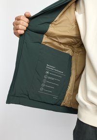 Green outer jacket with tan lining; features a pocket displaying care instructions and performance specifications; made from recycled materials.