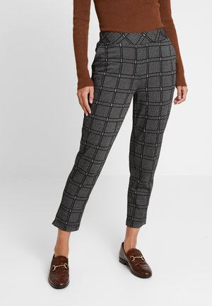 Vrouw draagt zwart-wit geruite broek tot aan de enkel, een bruin shirt met lange mouwen en donkerbruine loafers met gouden details.