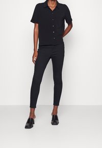 Personne portant une chemise noire à manches courtes avec boutons, un pantalon noir ajusté et des chaussures à talons noires sans lacets, sur un fond blanc uni.